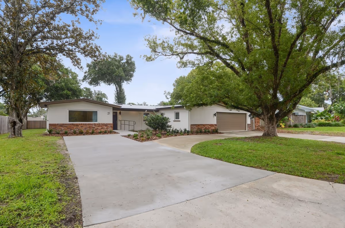 Front exterior of a single-story ranch-style home with brick accents, a wide concrete driveway, attached garage, and large trees on the lawn.