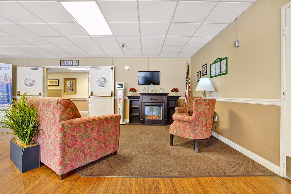 A cozy seating area in a senior living facility with two patterned armchairs and a small electric fireplace beneath a wall-mounted TV. There is a potted plant on the left, an American flag on the right, and a lamp on a side table. The walls are beige with white trim, and there is a sign above double doors that reads 'FAMILY'.