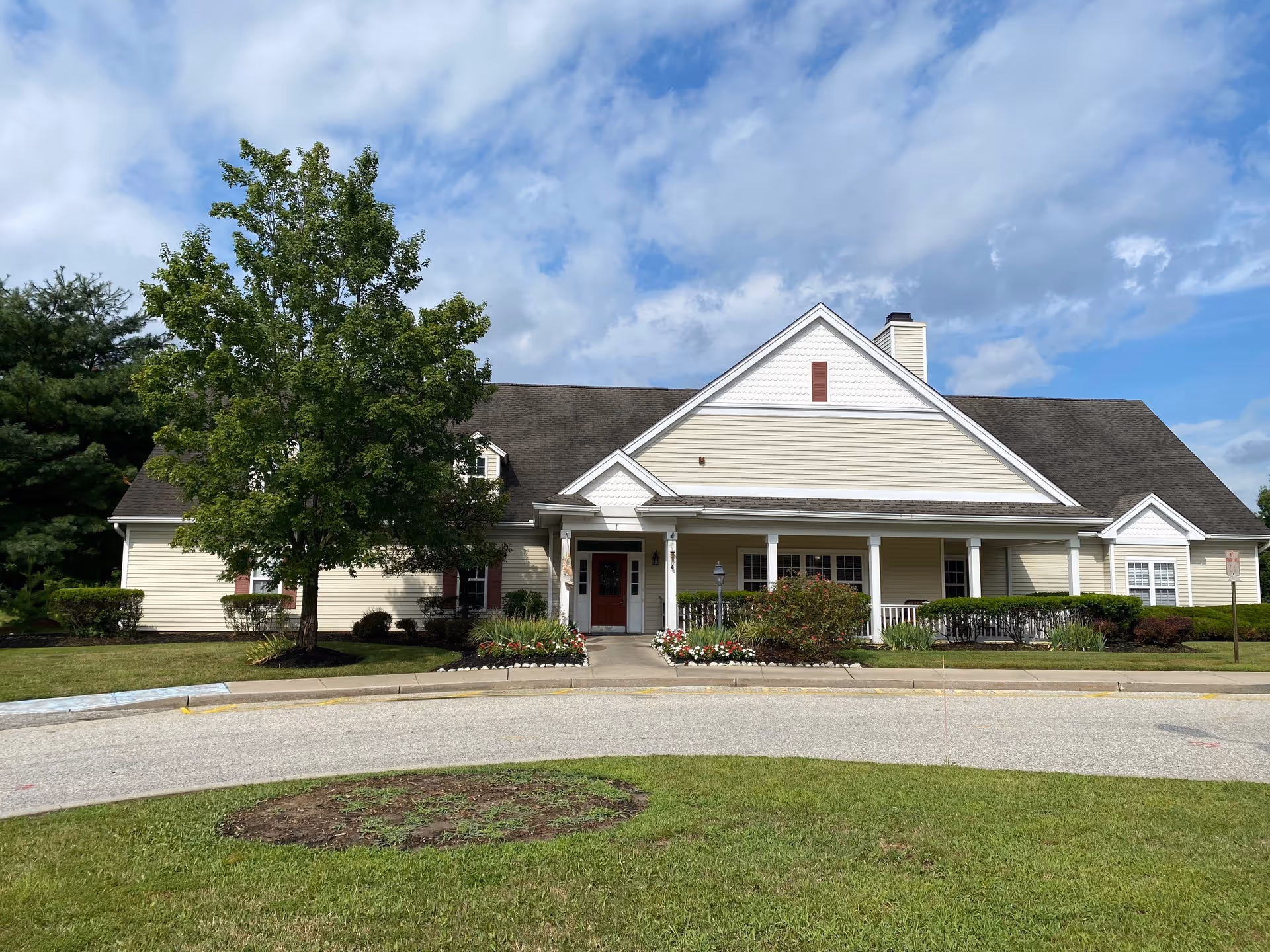 Exterior front view of a single-story senior living facility building with beige siding, a dark shingled roof, a covered porch with white columns, and a red front door. There is a tree and landscaped bushes and flowers in front of the building under a partly cloudy sky.