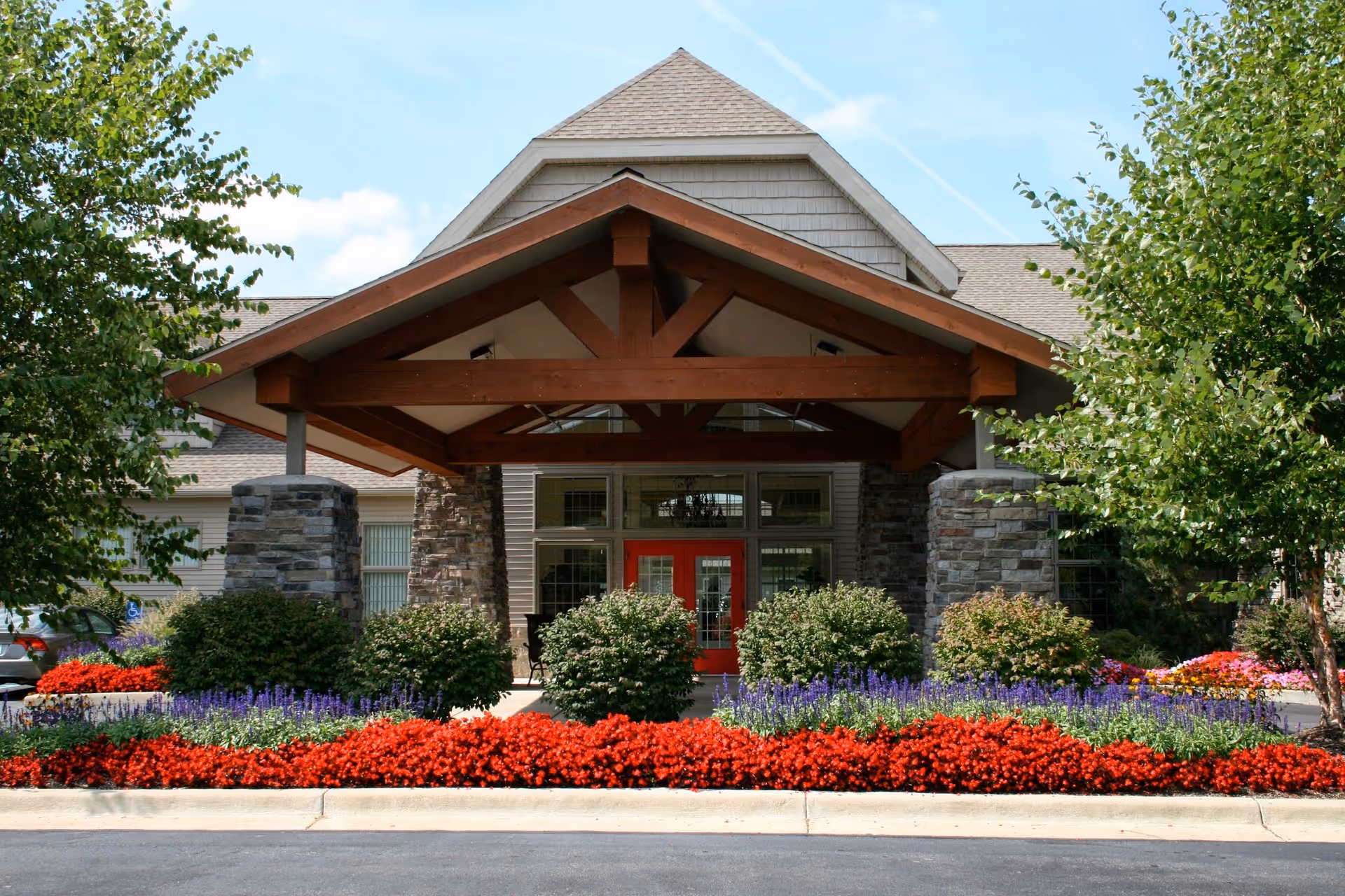 Exterior view of a senior living facility entrance with a wooden porte-cochère and vibrant landscaping.