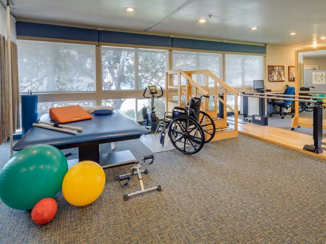 A bright rehabilitation room with a treatment table, exercise balls, a wheelchair, hand rails, parallel bars and practice stairs in front of large windows.