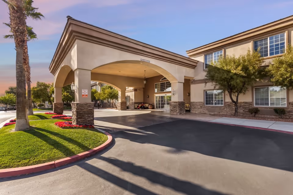 The front entrance and porte-cochère of a senior living building with palm trees, landscaped flower beds, and large windows.