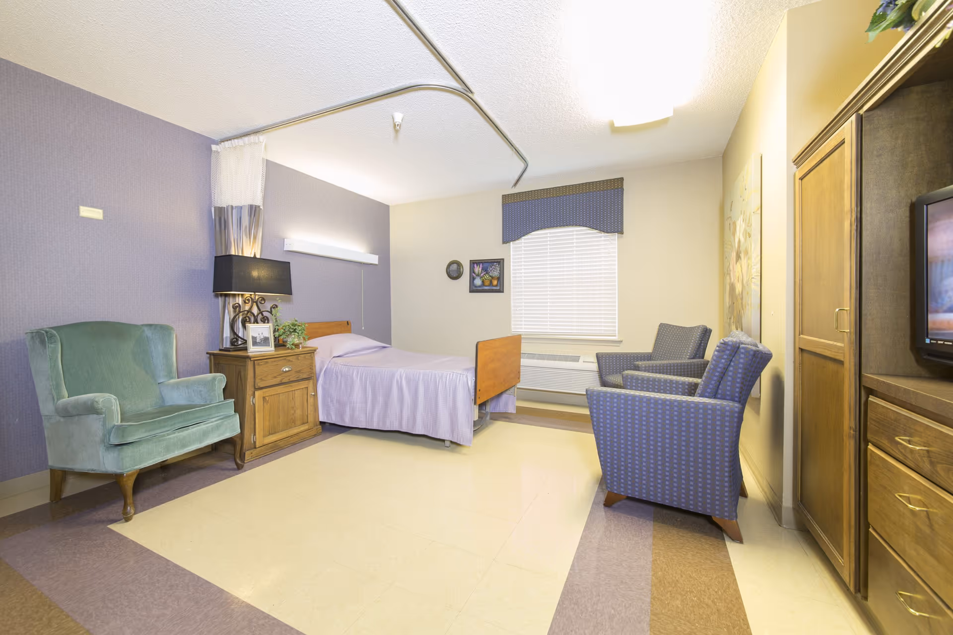 A nursing home bedroom with a single bed covered in a light purple bedspread, a wooden nightstand with a lamp and a small plant, a green armchair, two blue patterned armchairs, a window with blinds and a blue valance, and a wooden cabinet with a TV on top.
