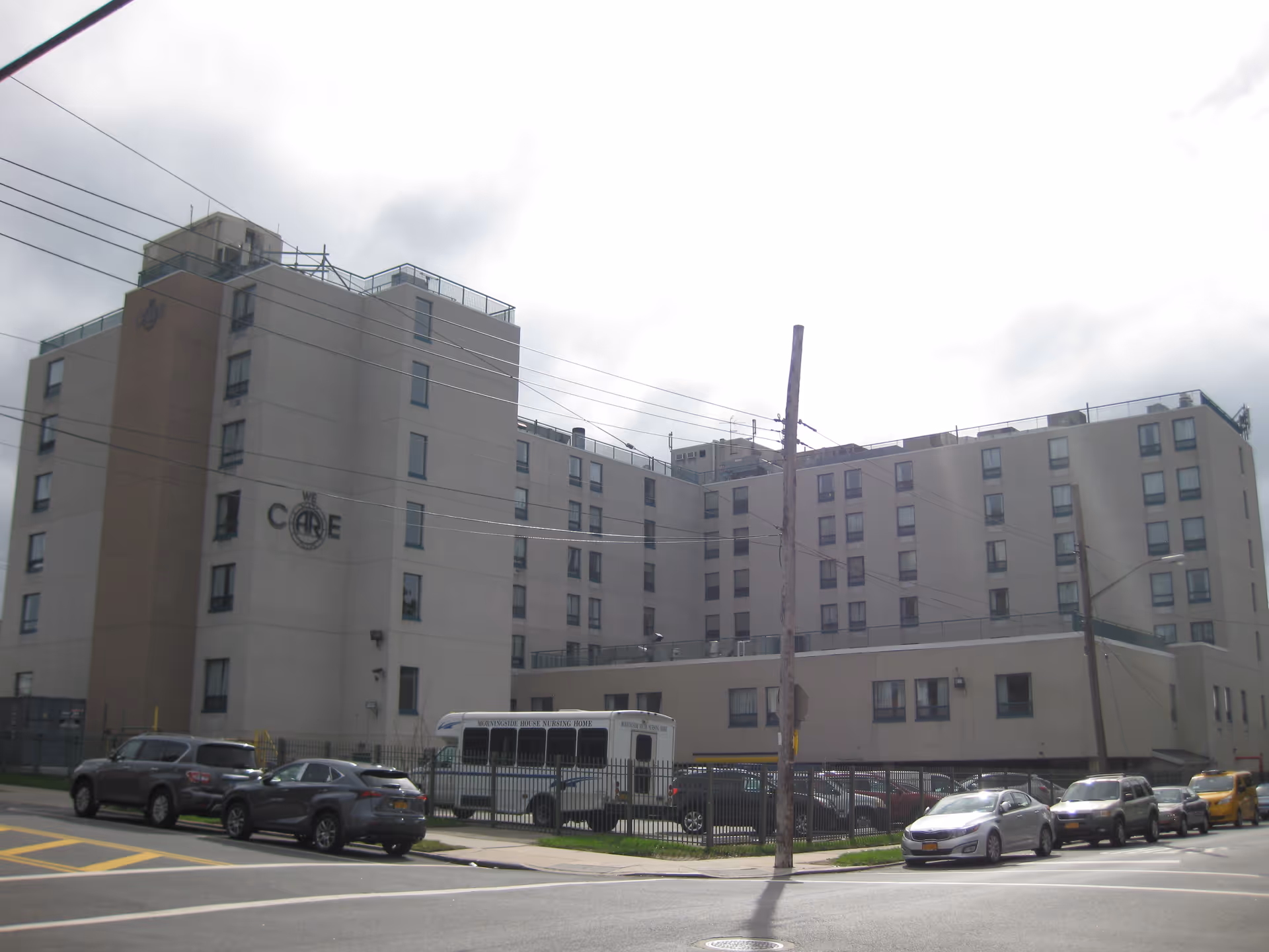 Exterior view of a multi-story senior care facility with a fenced parking lot and bus.
