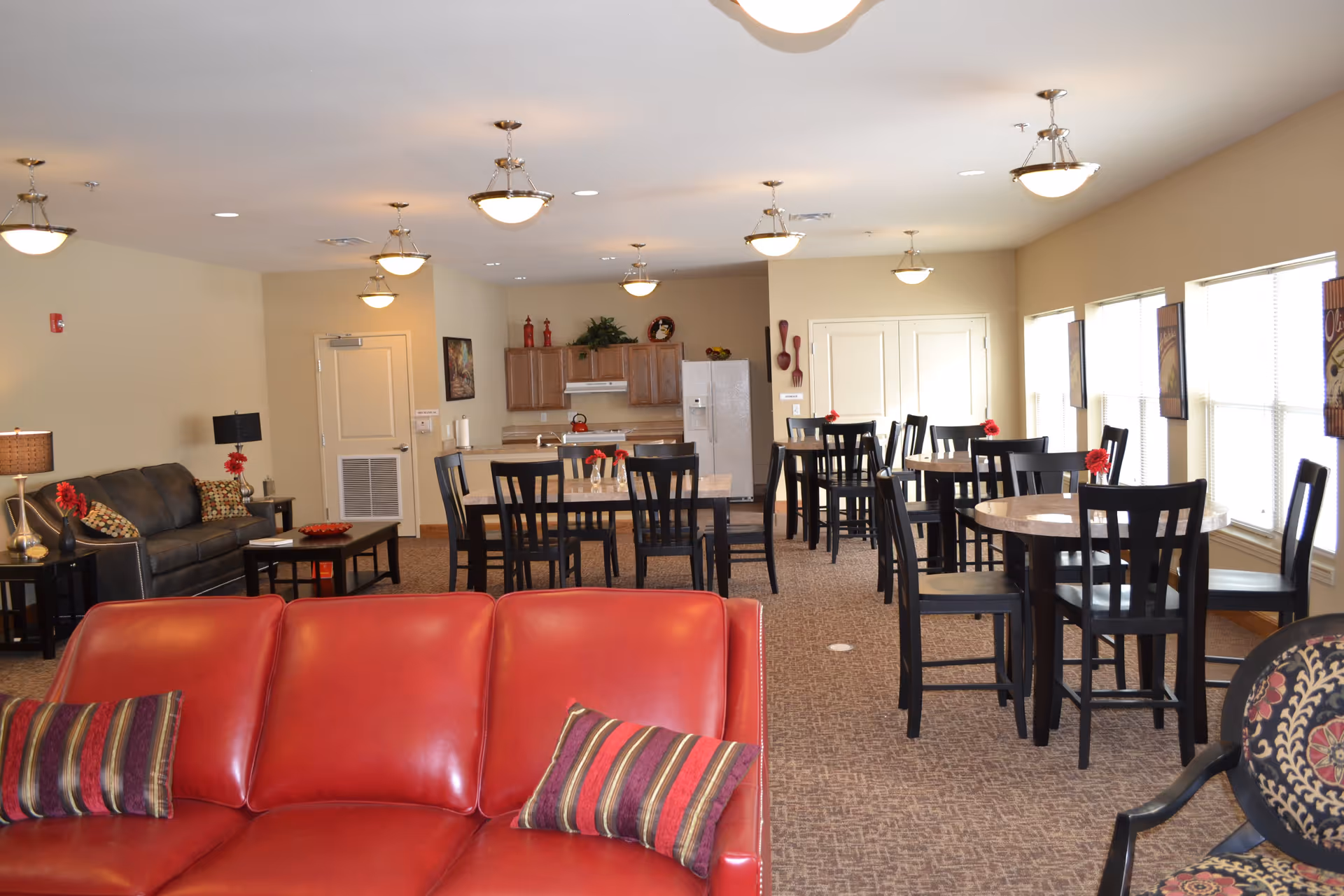 Open senior living common area with a red leather sofa in the foreground, multiple dining tables and chairs, lounge seating, and a kitchenette in the background.