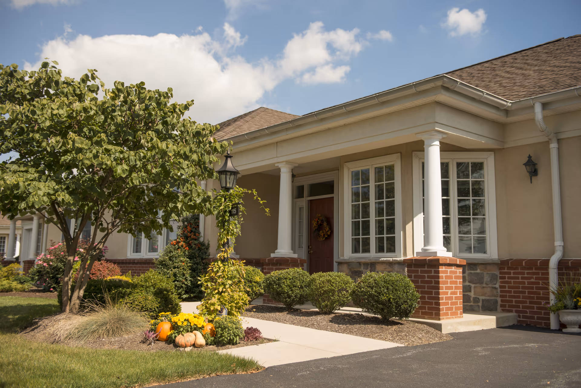 Exterior view of a single-story building with a covered porch supported by white columns, a red front door decorated with a wreath, large windows, and landscaped bushes and trees. There are pumpkins and flowers near the walkway leading to the entrance under a partly cloudy sky.