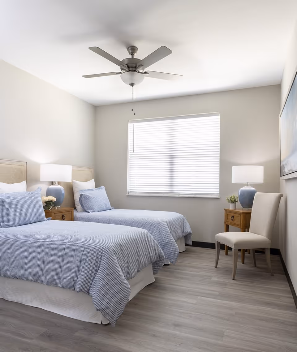 Bright bedroom with two twin beds, bedside tables and lamps, a chair, and a window with blinds.