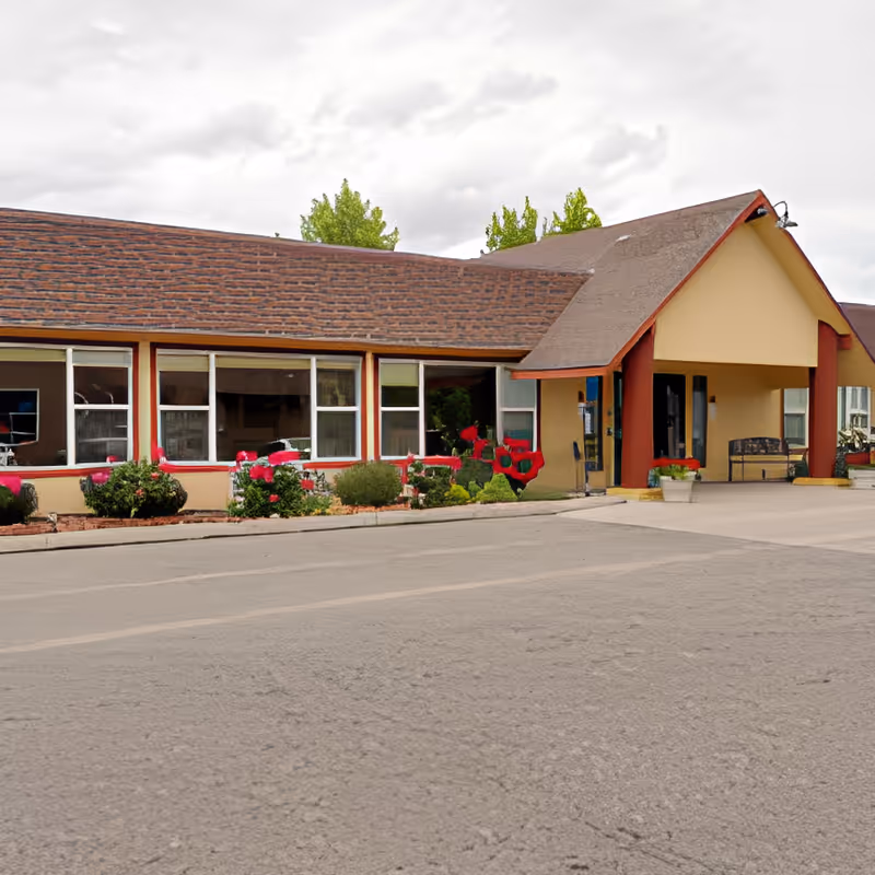 Exterior view of a single-story building with a sloped roof and large windows. The entrance has a covered porch with red pillars and a bench nearby. There are flower beds with red and pink flowers along the front of the building and some trees visible in the background under a cloudy sky.