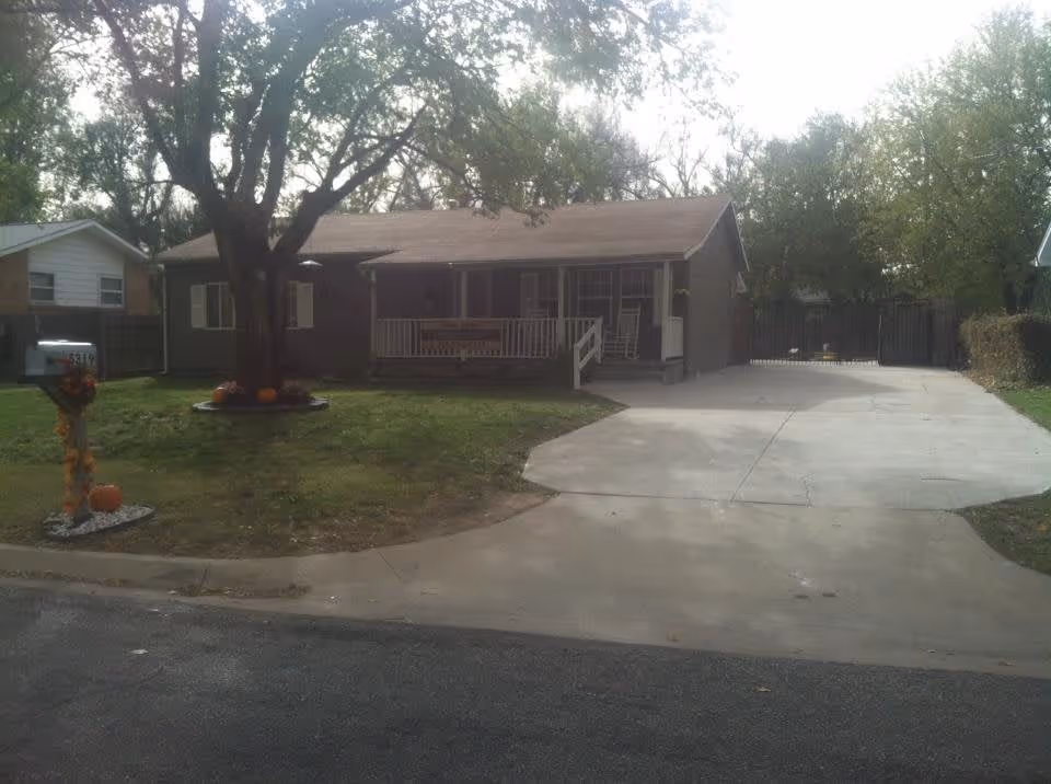 Single-story house with a front porch, a large tree in the yard, and a concrete driveway. The mailbox is decorated with autumn-themed items including pumpkins. The house is surrounded by other homes and trees.