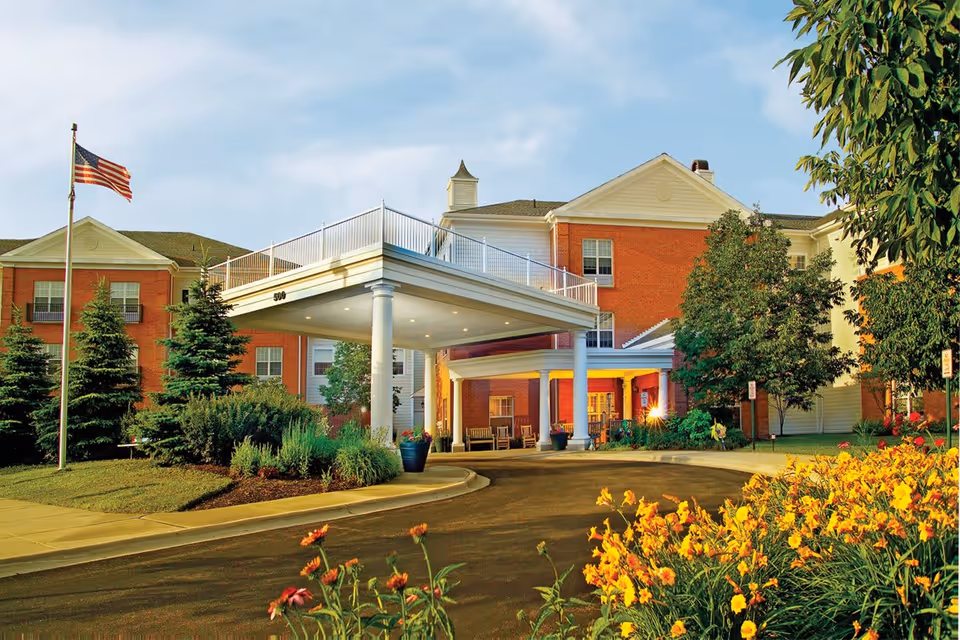 Exterior view of Belmont Village Senior Living Buffalo Grove building with a covered entrance, American flag on a flagpole, landscaped greenery, and yellow flowers in the foreground under a partly cloudy sky.