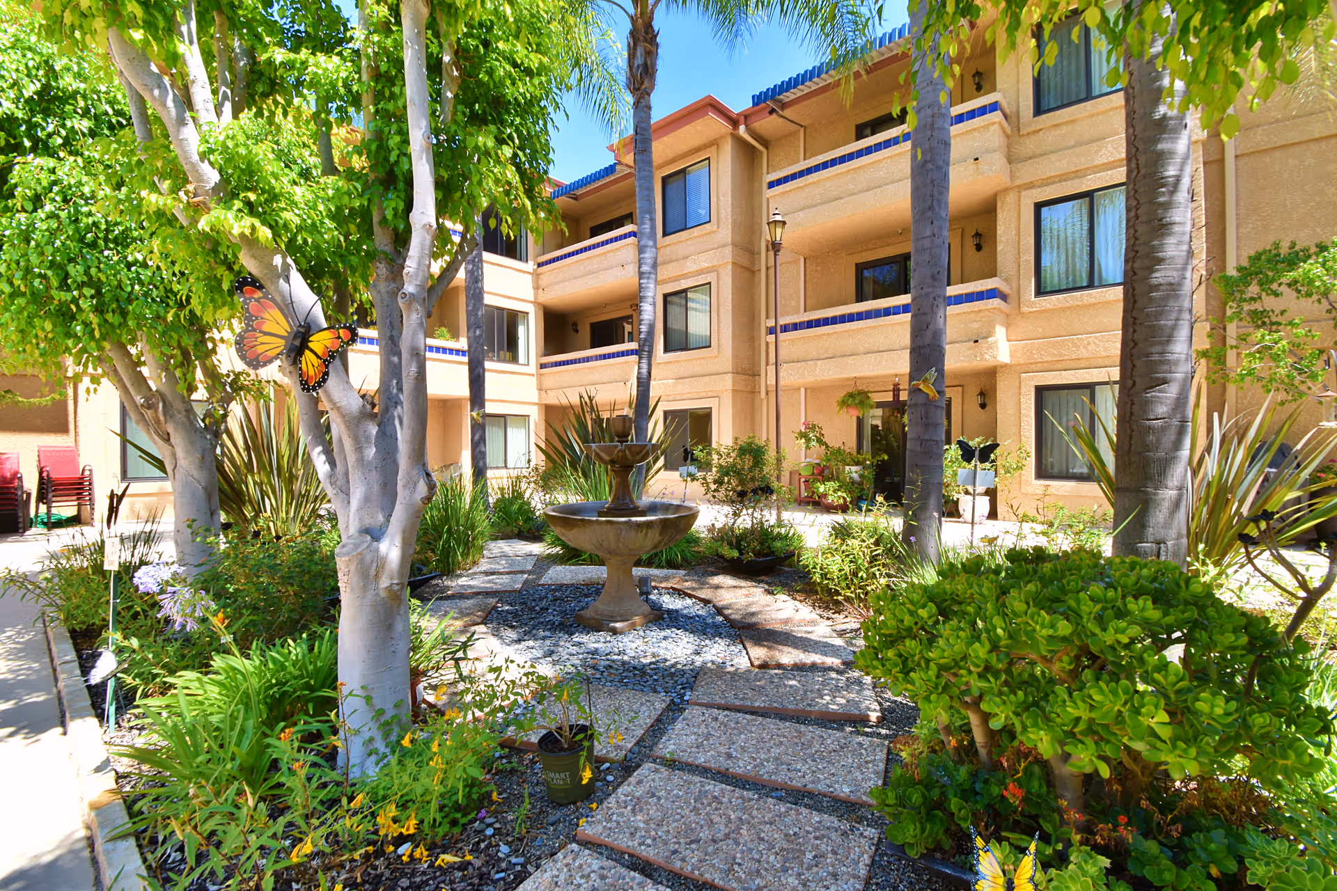 Sunny courtyard at Freedom Village featuring a fountain, lush plants, and a beige multi-story building.