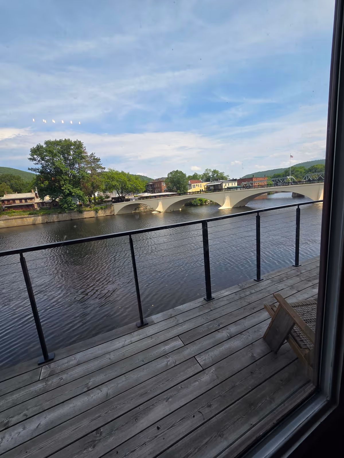 Outdoor wooden deck overlooking a river, arched bridge, trees, and colorful buildings across the water.