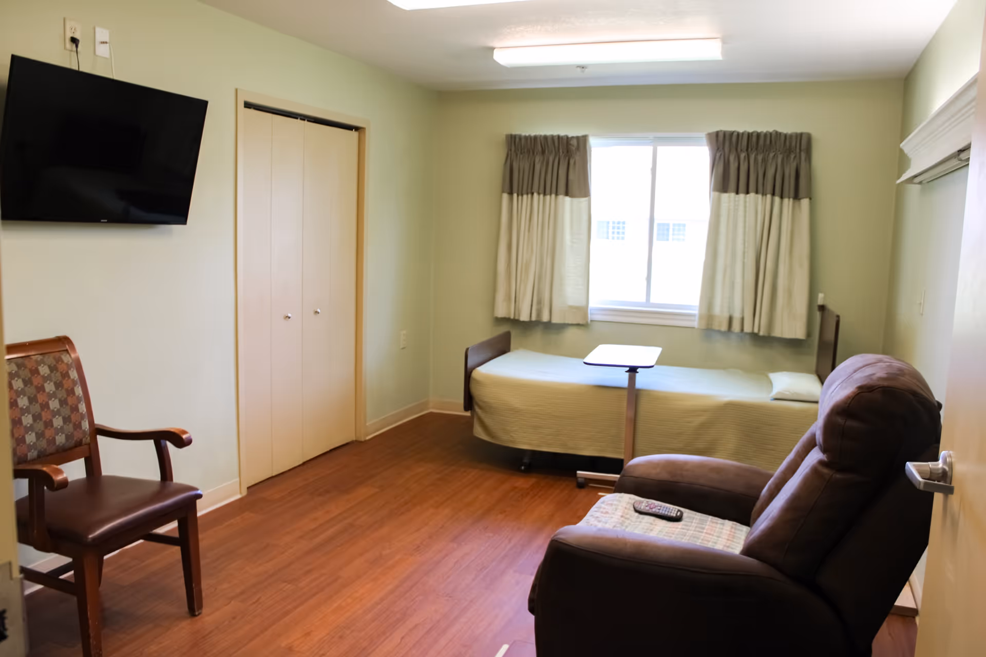 A small resident bedroom with bed, recliner, TV, closet, and a window with curtains.