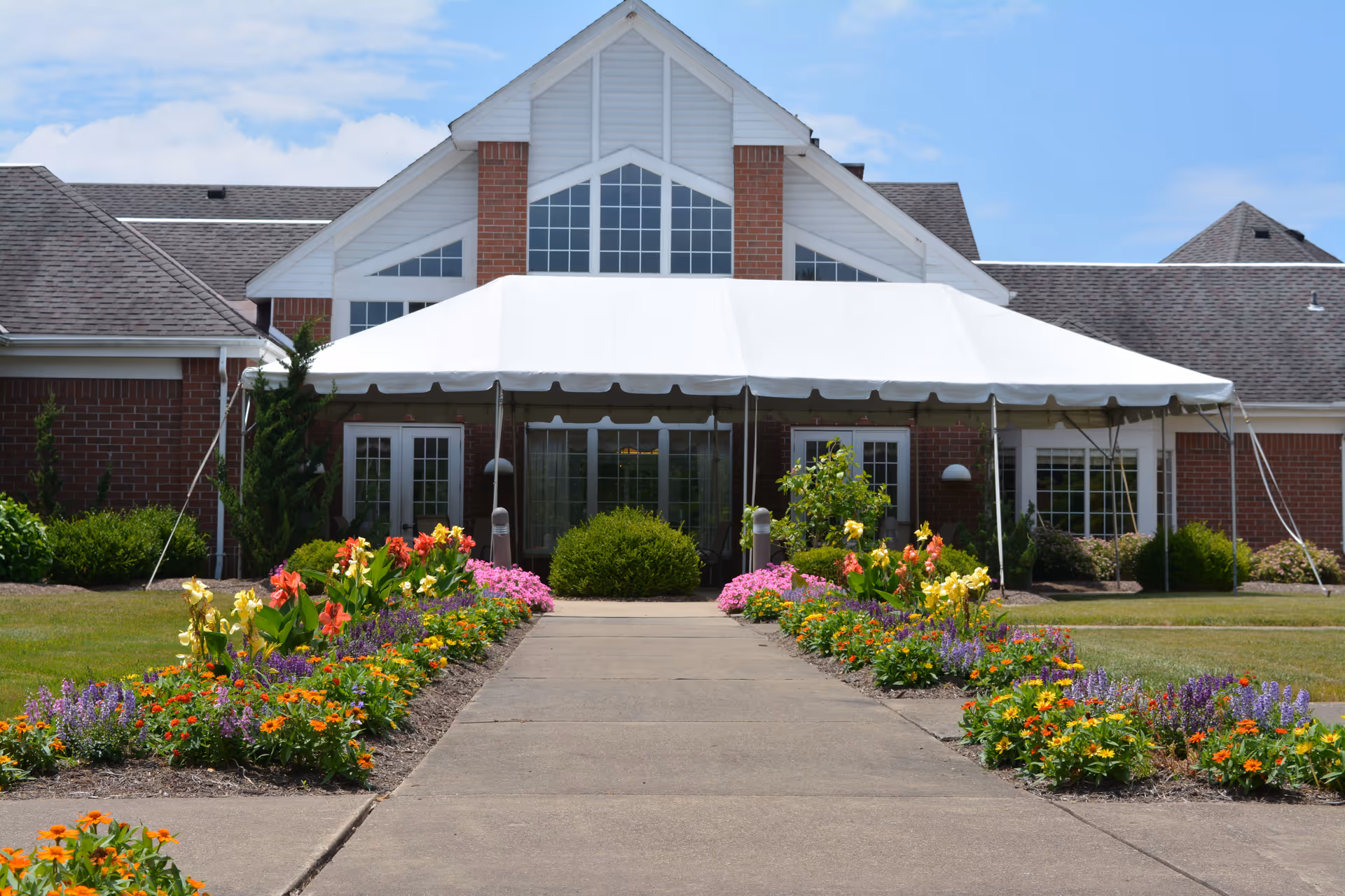 Front exterior view of a brick building with a large white tent covering the entrance. The walkway leading to the entrance is lined with colorful flower beds on both sides. The building has large windows and a peaked roof under a blue sky with some clouds.