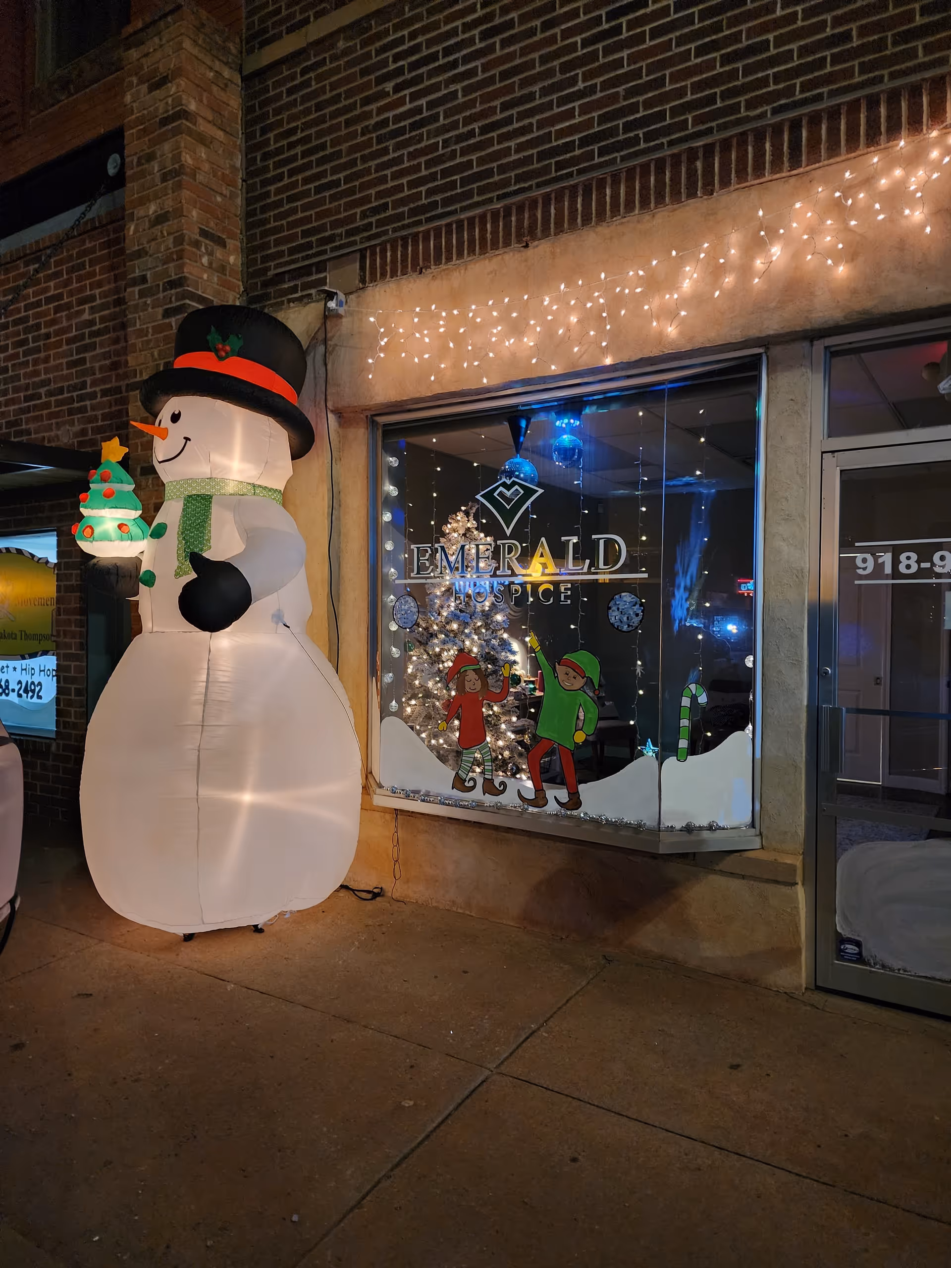 Nighttime storefront window displays Emerald Hospice with holiday decor and inflatable snowman.