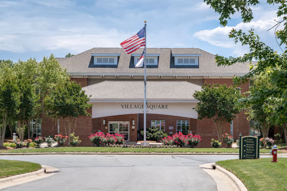 Front entrance of a brick senior living building labeled 'Village Square' with an American flag, landscaped flowerbeds, and a driveway.