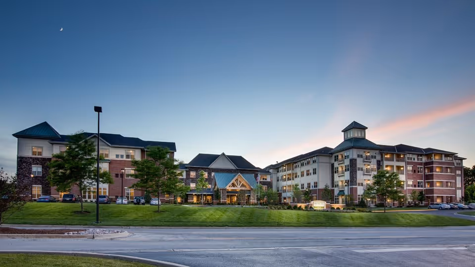 Exterior view of Deer Park Village senior living facility at dusk, showing a large multi-story building with a mix of brick and light-colored siding, a peaked entrance with a blue roof, several trees, parked cars, and a clear sky with a crescent moon.