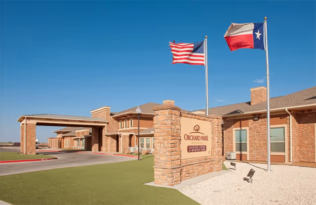 Exterior view of Orchard Park of Permian Basin senior living facility with brick building and flags.