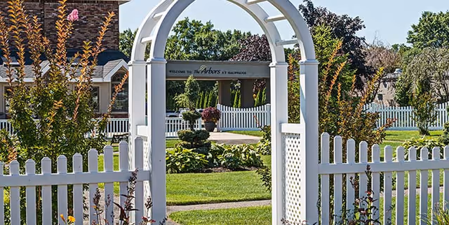 White archway entry with picket fence leads to The Arbors at Hauppauge entrance.