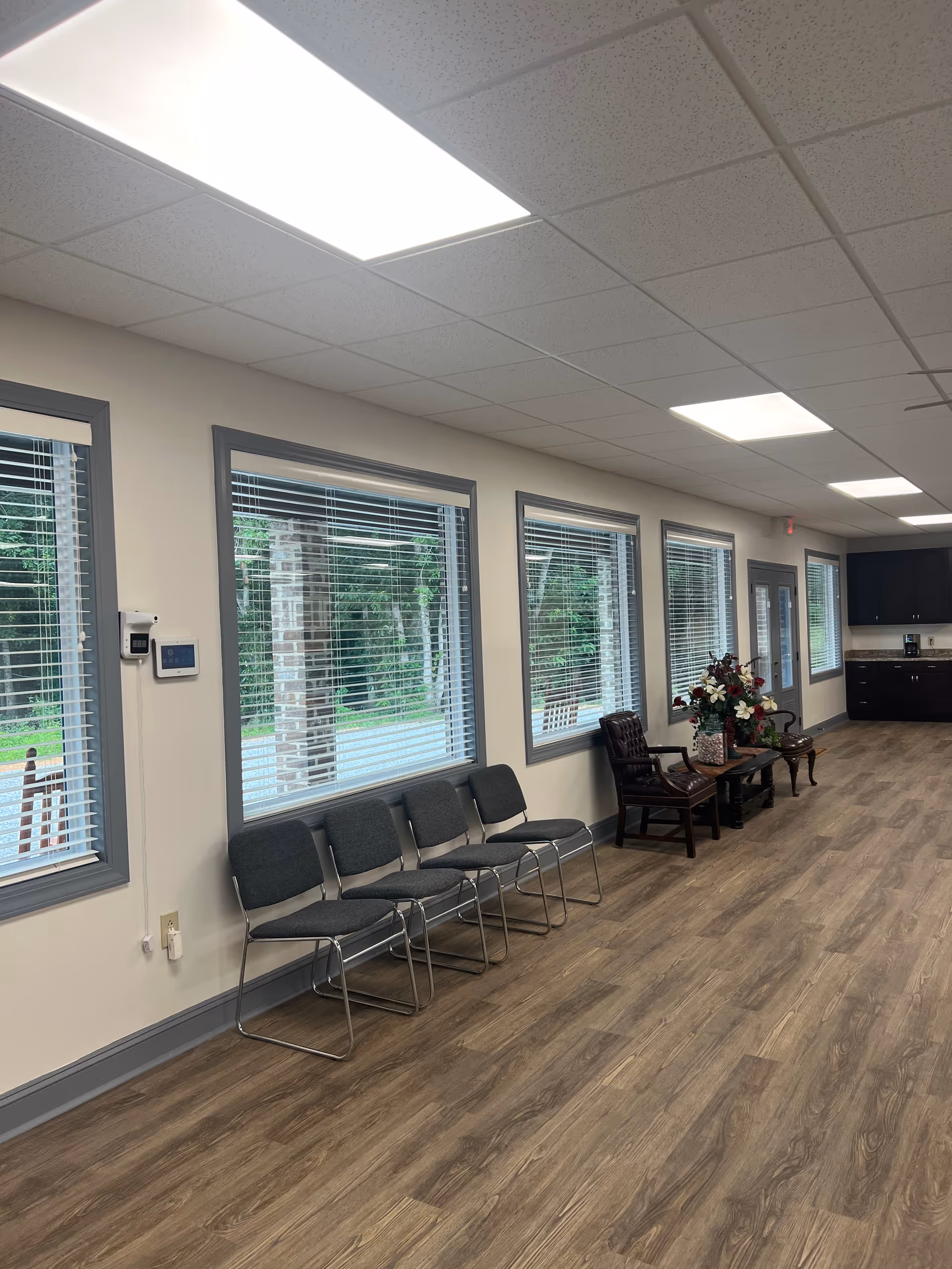 Interior waiting area with row of gray chairs along windows and a kitchenette in back.