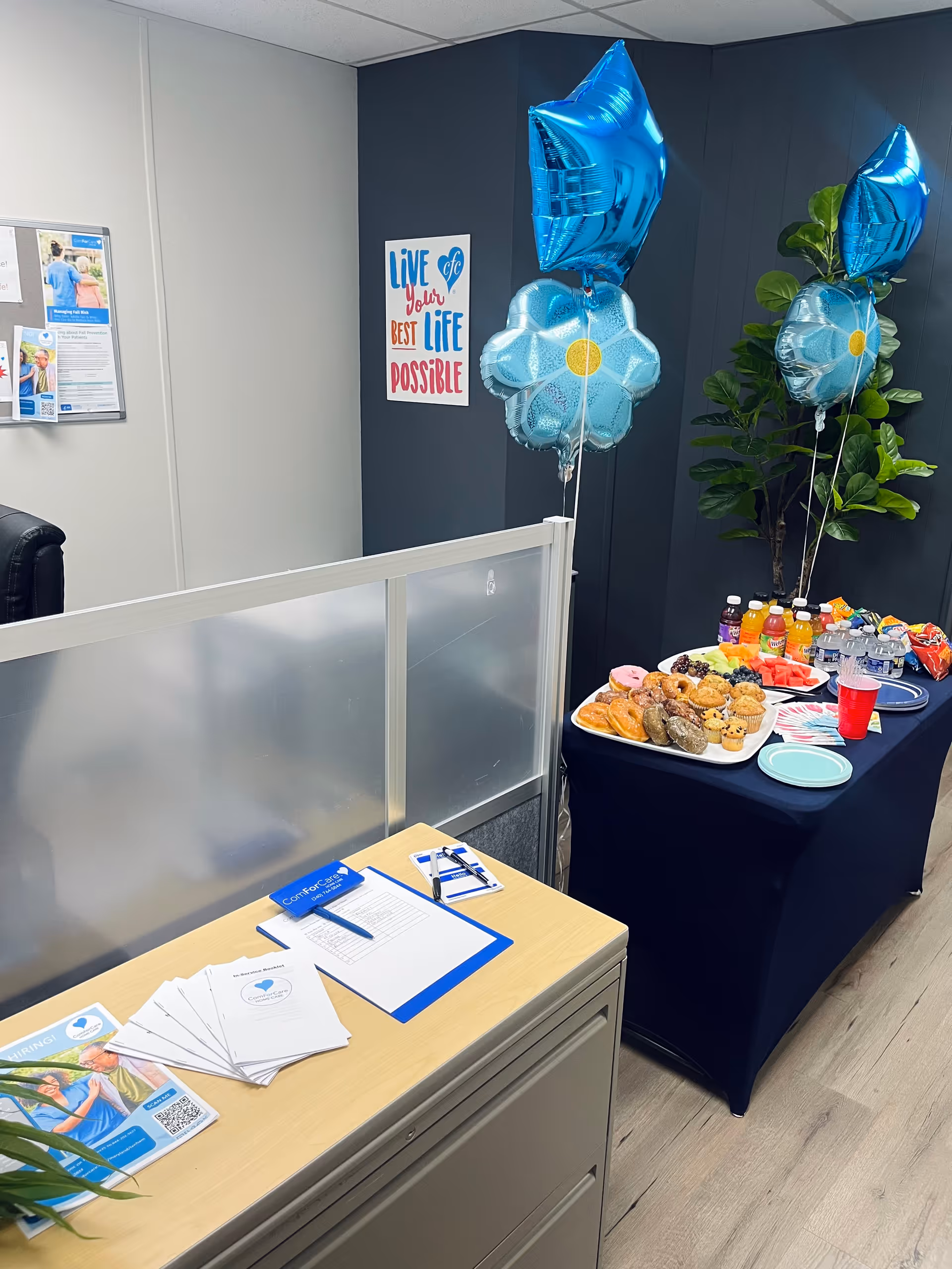 Interior corner of a senior care facility with a refreshment table, balloons, plants, and brochures.