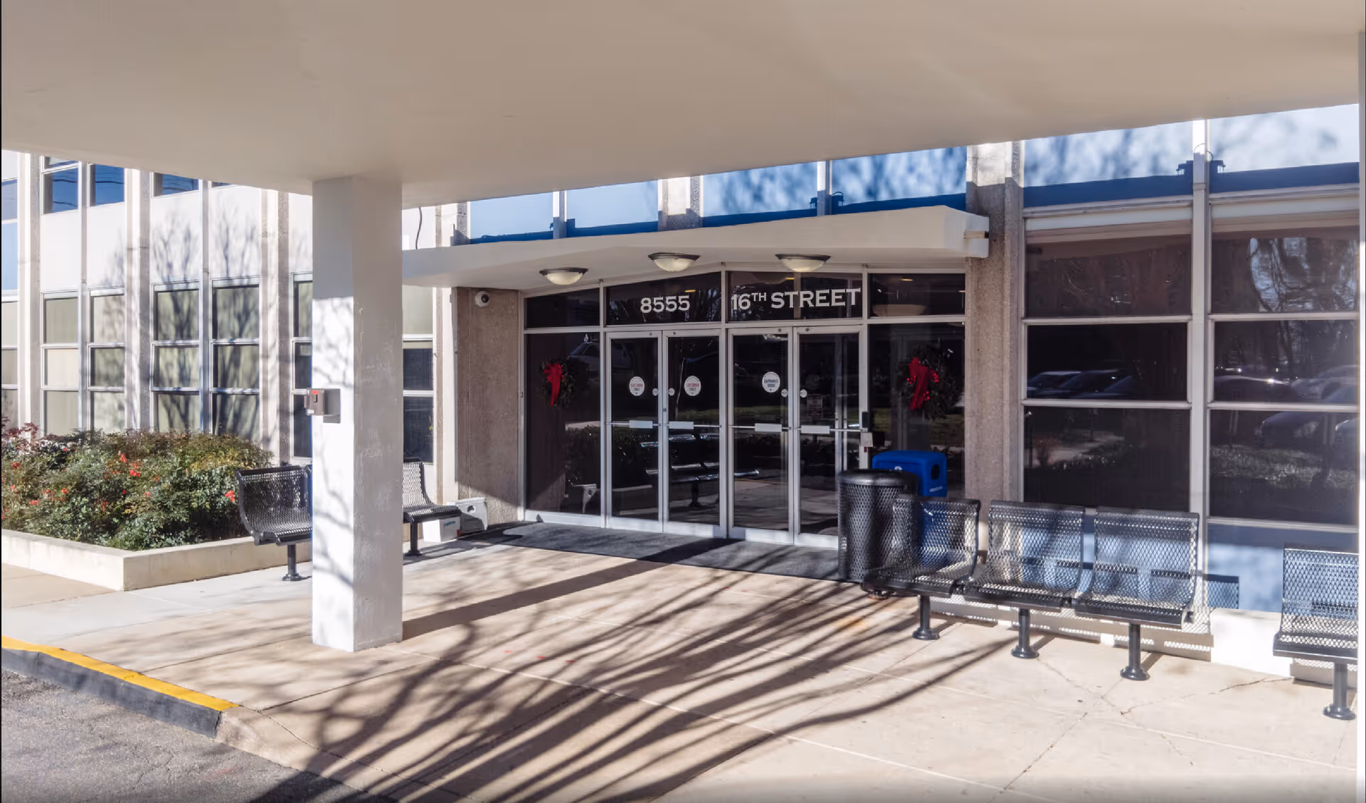 Exterior view of a senior living facility entrance with glass doors, benches, and a shaded walkway.