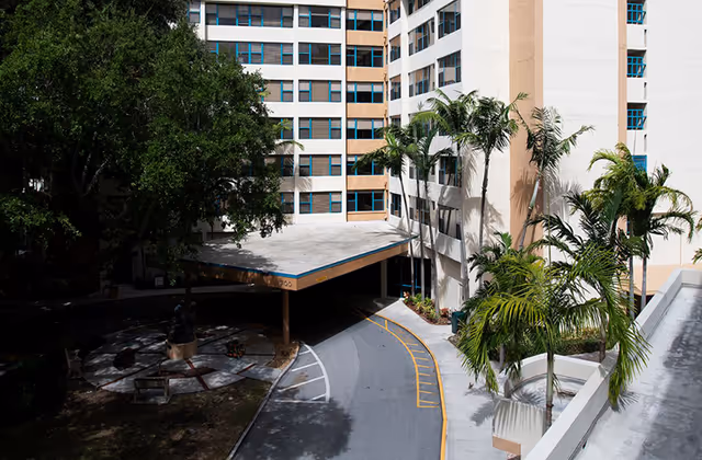 Exterior view of Irving Cypen Tower showing a multi-story building with beige and white walls and blue window frames. There is a covered driveway entrance with a curved road leading up to it, surrounded by palm trees and other greenery.