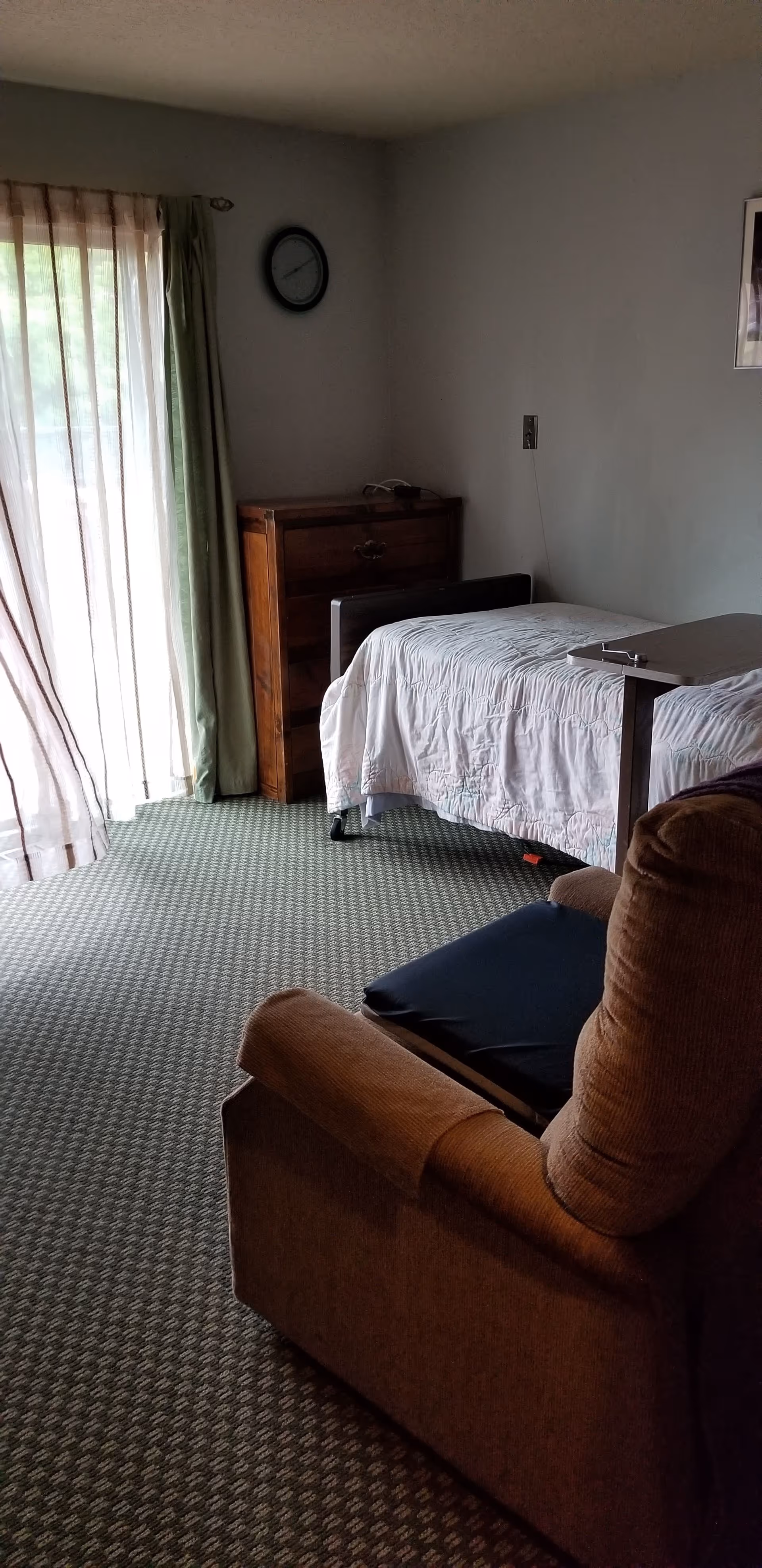 A small bedroom with a single bed covered in a white quilt, a wooden chest of drawers against the wall, a clock above it, and a brown recliner chair in the foreground. Light filters through sheer curtains covering a sliding glass door on the left side.