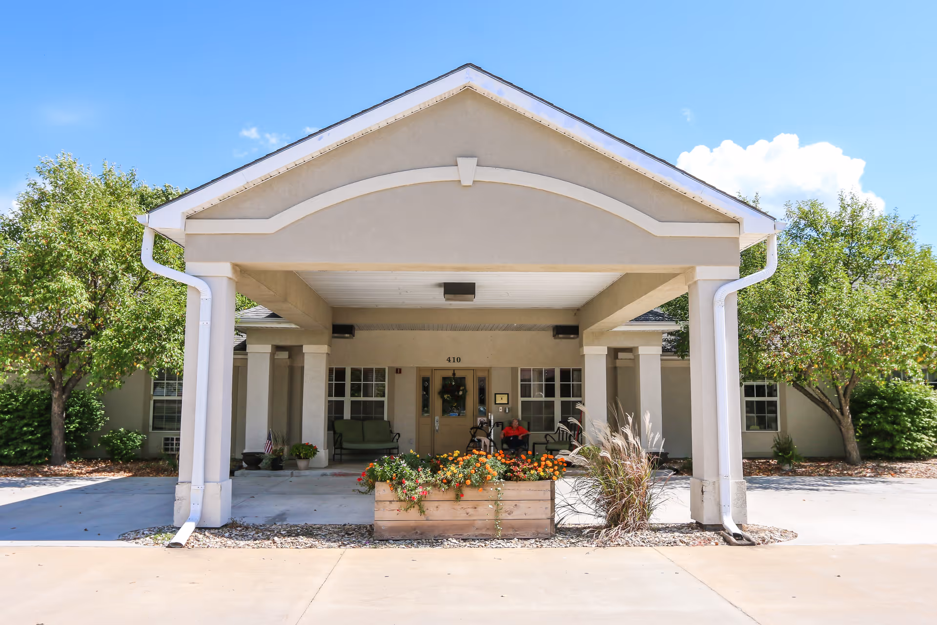 Front entrance of a senior living facility with a covered driveway supported by columns. There are flower beds with colorful flowers in front, trees on both sides, and a person sitting on a bench near the entrance door under the covered area.