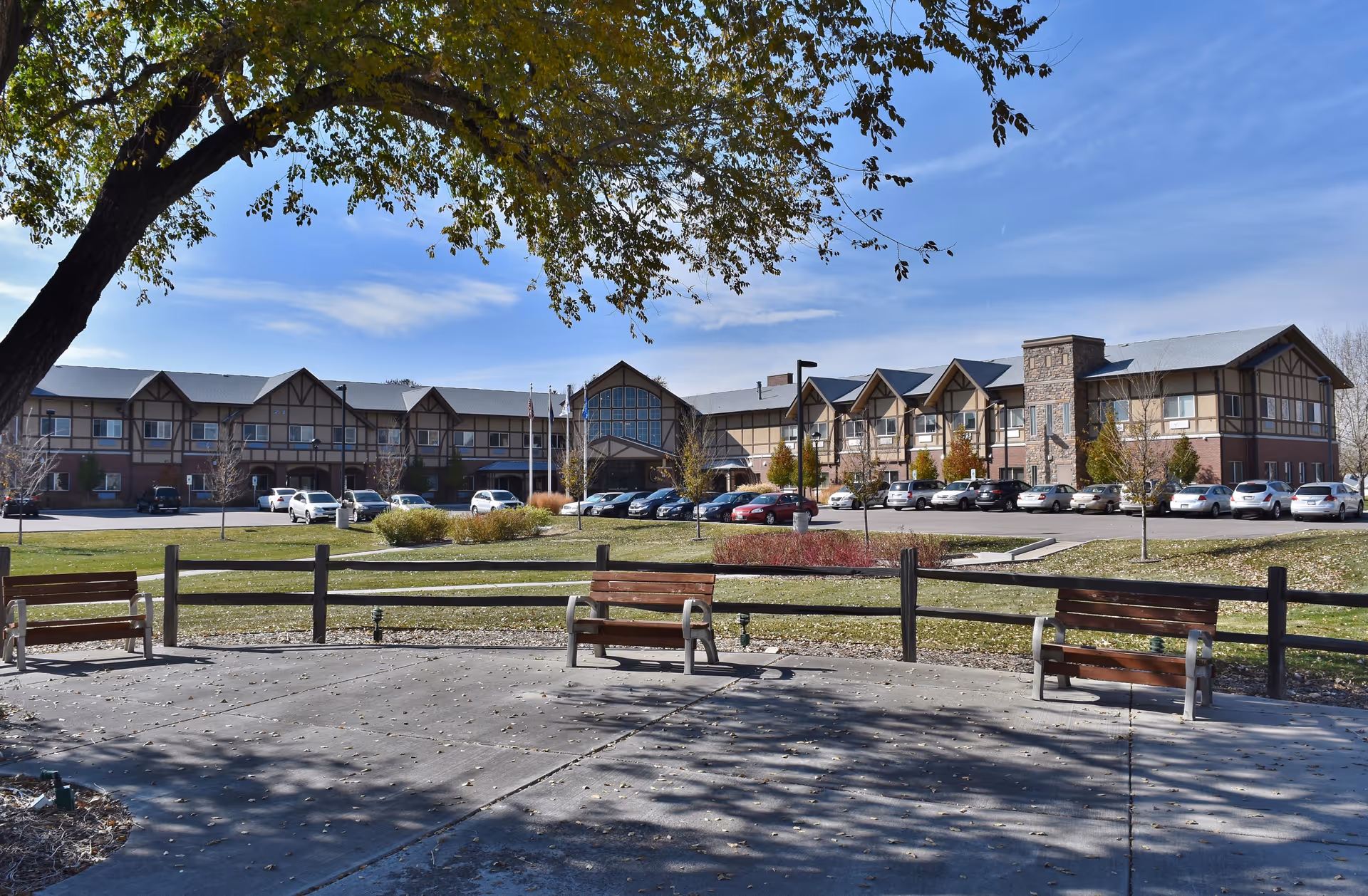 Exterior view of Eastern Star Masonic Retirement Campus building with a parking lot in front. There are several cars parked and a grassy area with benches and a tree in the foreground under a clear blue sky.