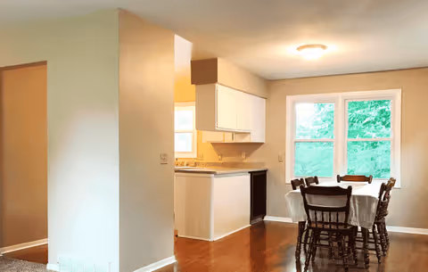 Open dining and kitchen area in a senior living home, with a table and windows.