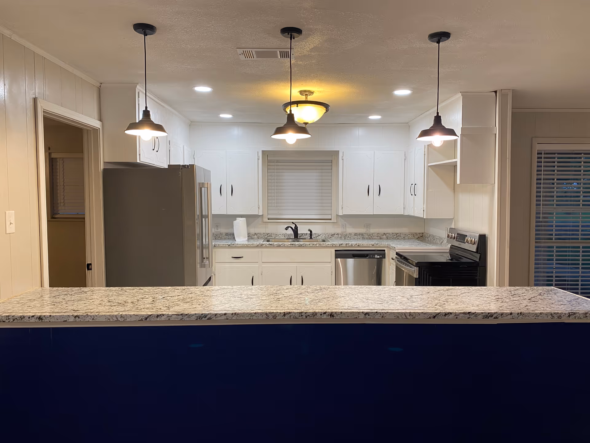 Modern kitchen with white cabinets, granite countertops, stainless steel refrigerator, dishwasher, and stove. Three pendant lights hang over a granite countertop bar area. A window with blinds is above the sink, and there is a door to the left and a window with blinds to the right.