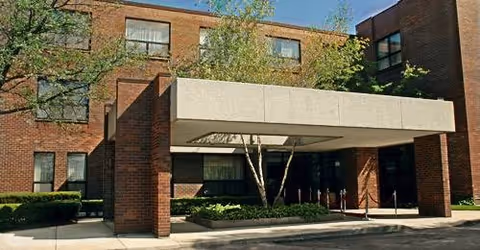 Brick senior living facility with a covered entrance and trimmed shrubs.