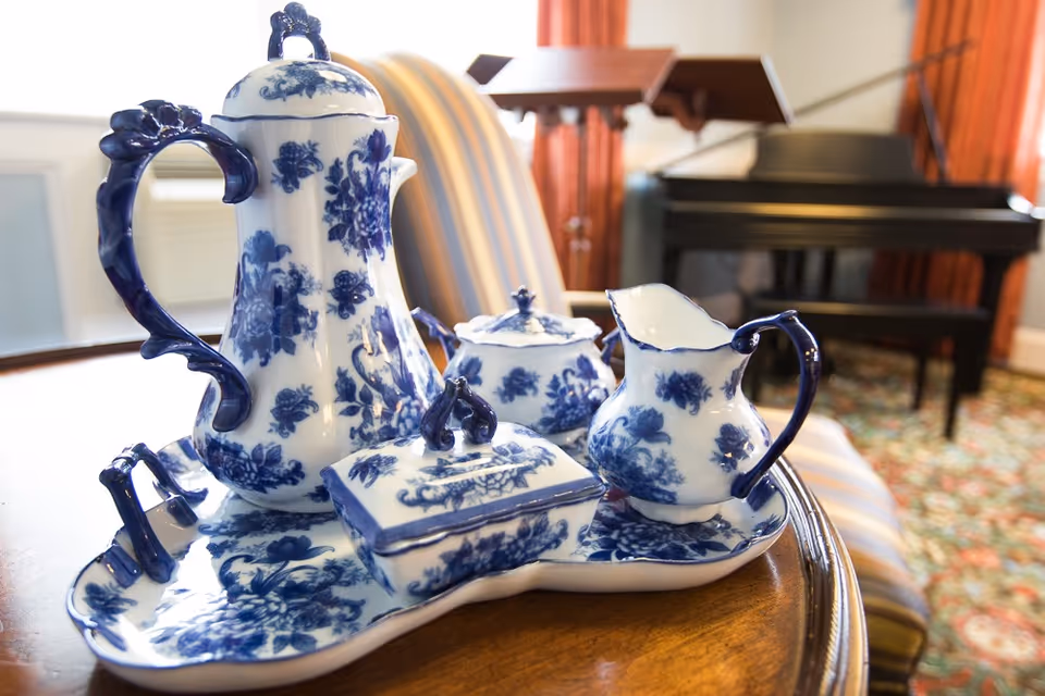 A close-up view of a blue and white floral porcelain tea set on a wooden table, with a striped armchair and a black grand piano in the background.