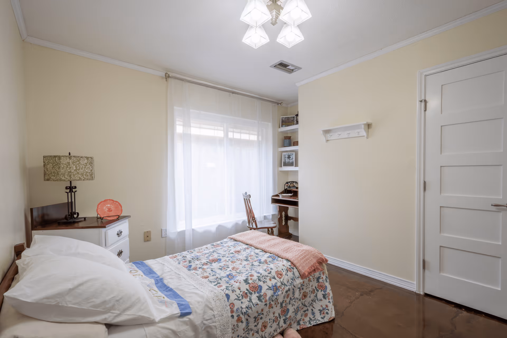 Cozy bedroom with floral quilt, white furniture, desk, shelves, and sheer-windowed light.