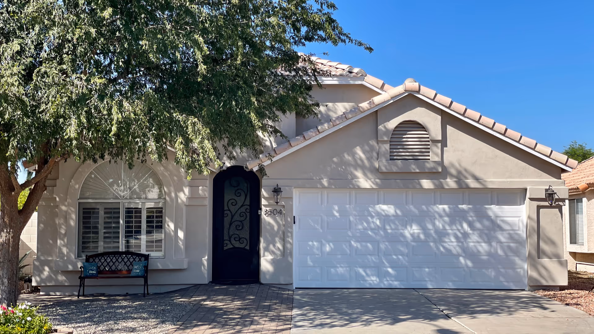 Front exterior view of a single-story house with a light beige facade, a large white garage door, an arched black front door with decorative ironwork, a window with white shutters, a bench with two blue pillows, and a large tree providing shade in the front yard under a clear blue sky.