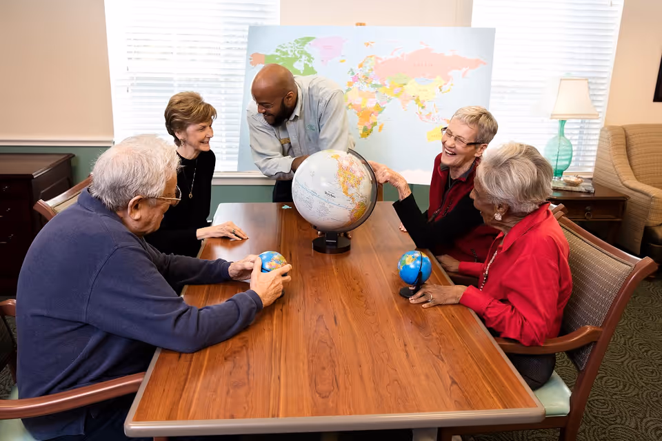A group of four elderly individuals and a staff member gathered around a wooden table in a well-lit room. They are engaged with three globes on the table, smiling and interacting. Behind them is a large world map on the wall, and the room has windows with blinds, a lamp, and comfortable chairs.