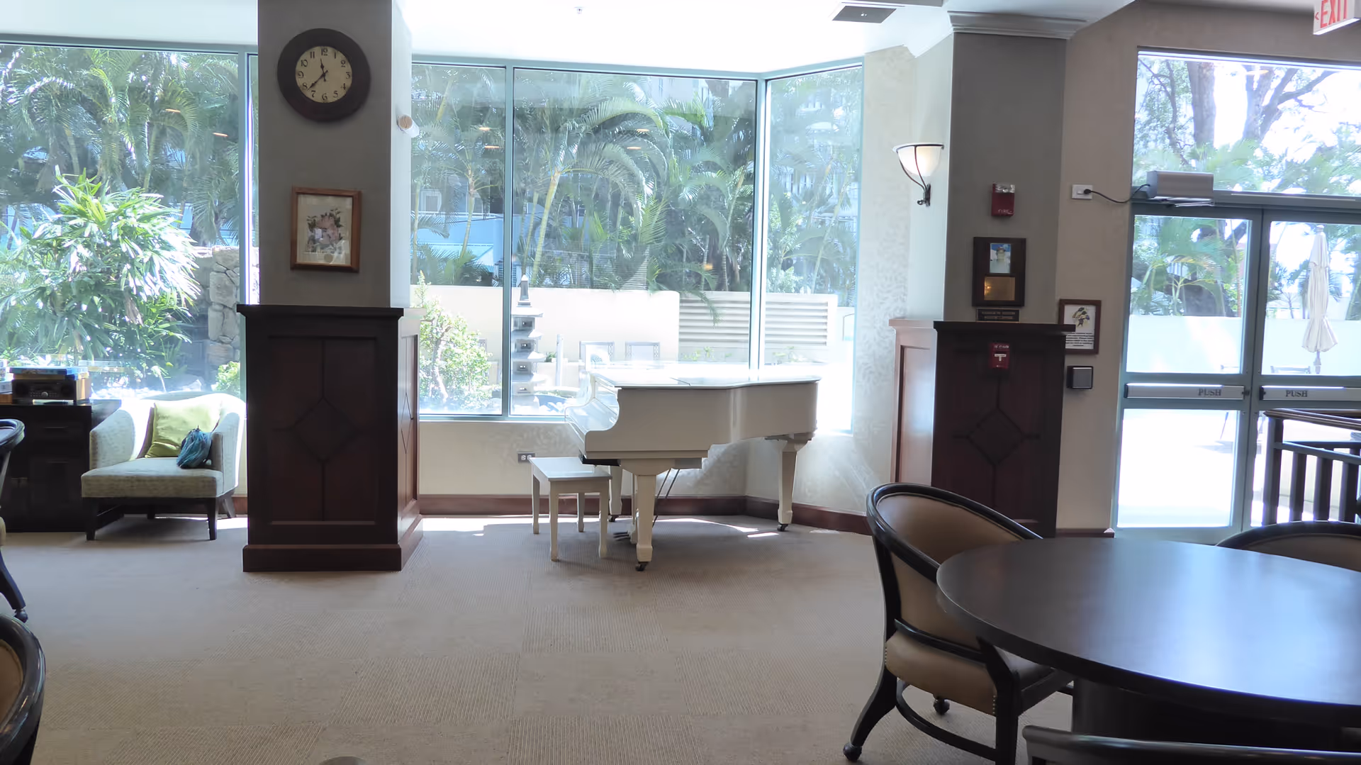 Sunlit senior living lobby with a white piano, round table, chairs, and large windows.