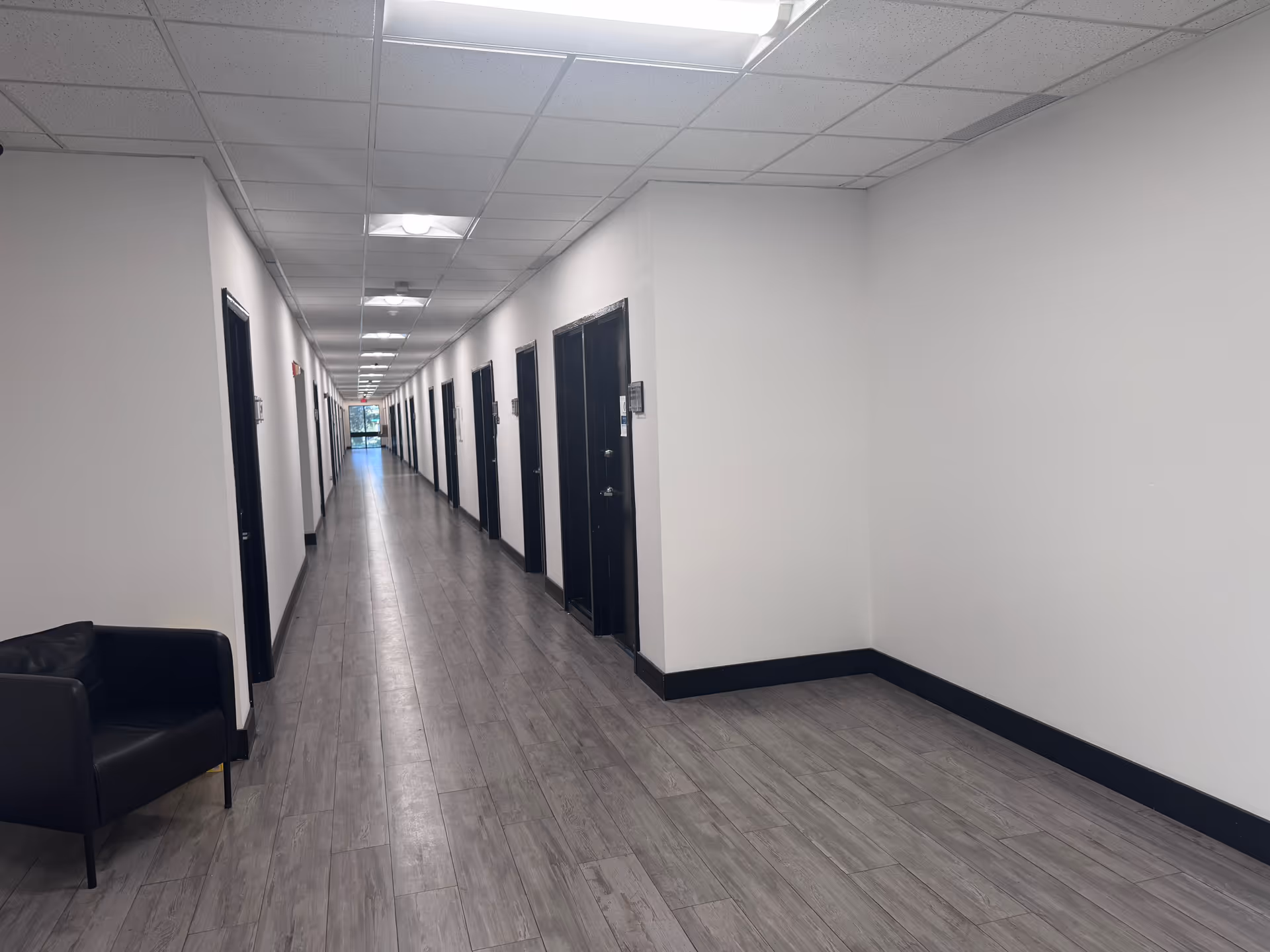Long white hallway in a senior living facility with doors, ceiling lights, and a chair.