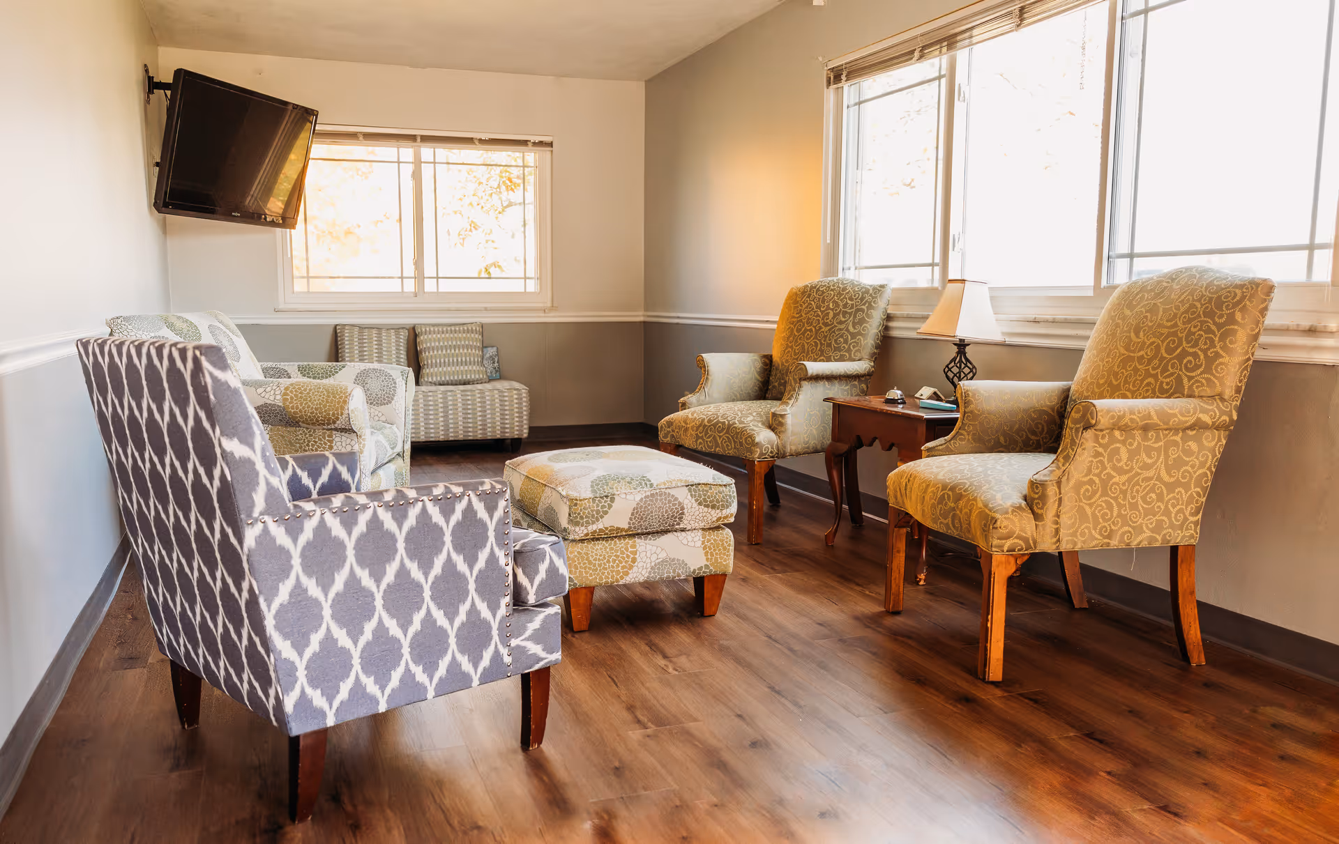 A cozy sitting area in a nursing home with patterned armchairs, an ottoman, a small wooden side table with a lamp, and a wall-mounted flat-screen TV. The room has large windows letting in natural light and wooden flooring.