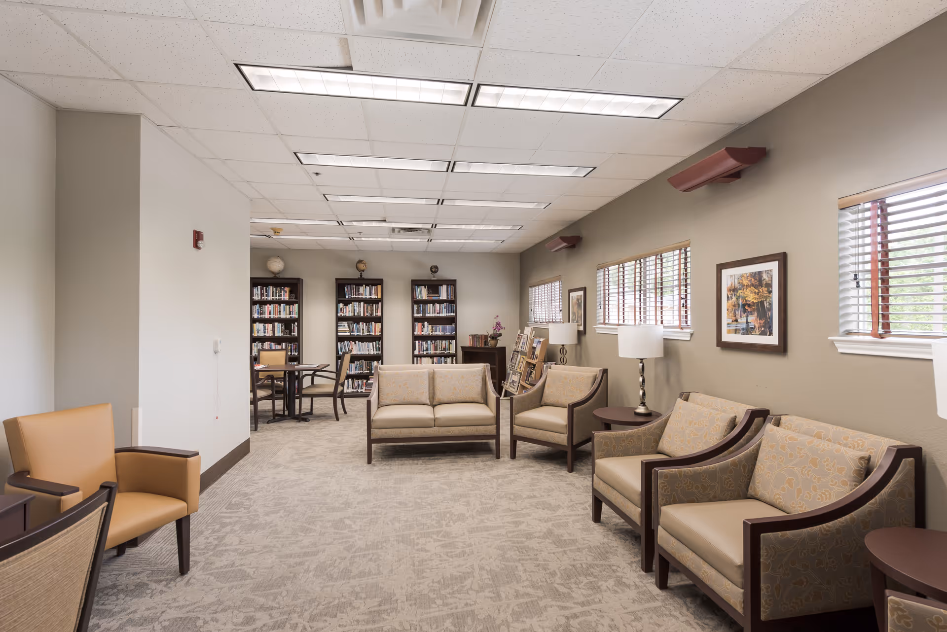 A well-lit common area in a senior living facility featuring multiple cushioned chairs and sofas arranged around small tables. The room has beige walls with framed artwork, windows with blinds, and a carpeted floor. In the background, there are three tall bookshelves filled with books and a small table with chairs for reading or activities.