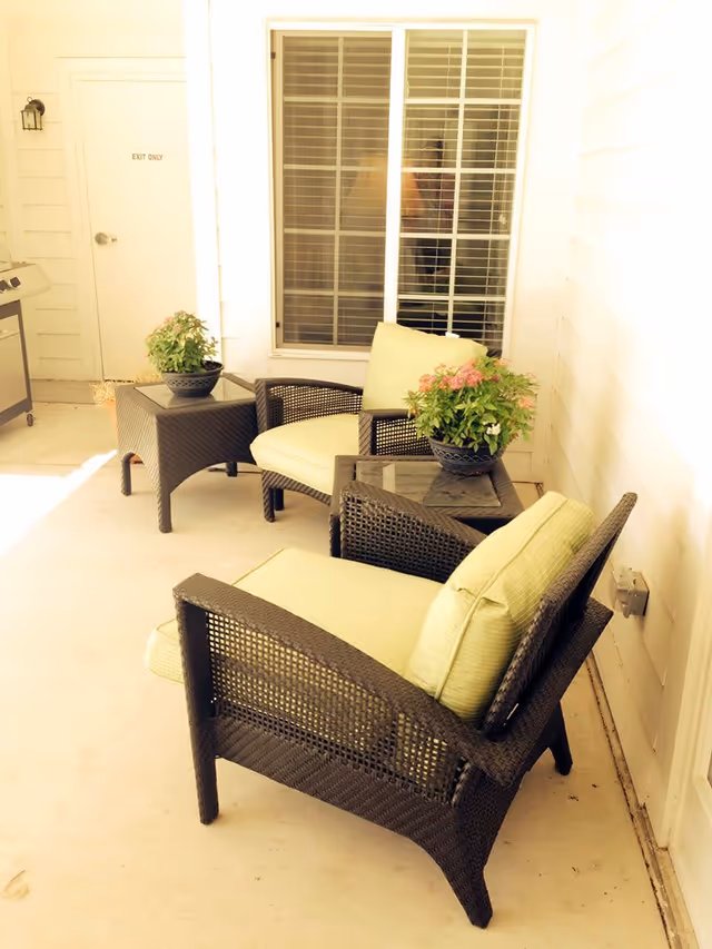 Outdoor patio area with two wicker chairs with light green cushions, two matching wicker side tables with potted plants, a window with blinds, and a door labeled 'EXIT ONLY'.