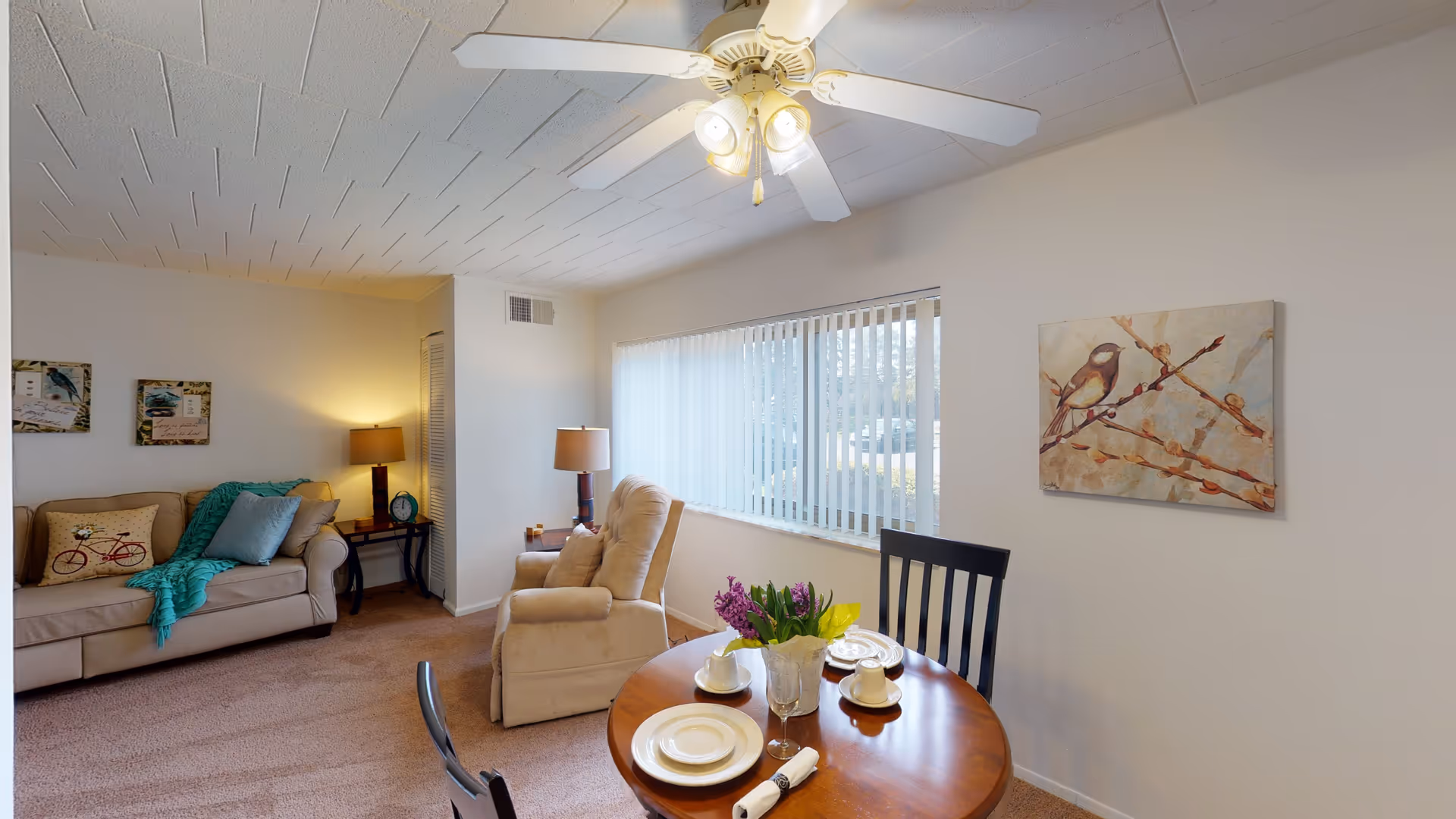 Bright senior living room with a sofa, recliner, small round dining table set with dishes, ceiling fan, and a window with vertical blinds.