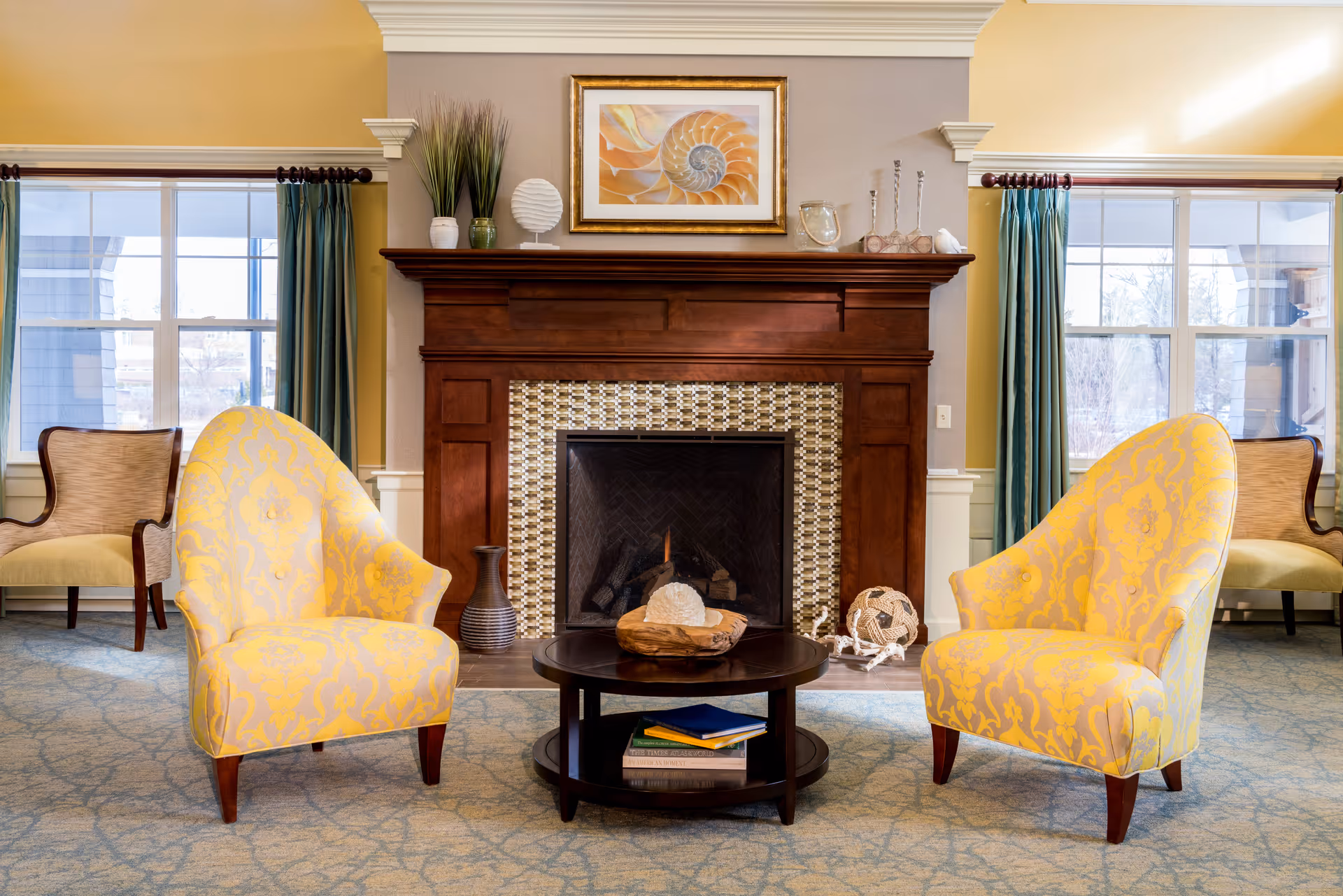 A cozy living room area featuring two yellow patterned armchairs facing a dark wooden coffee table with books and decorative items. Behind the table is a fireplace with a wooden mantel adorned with vases, a framed seashell artwork, and other decorative objects. Large windows with teal curtains flank the fireplace, allowing natural light into the room.