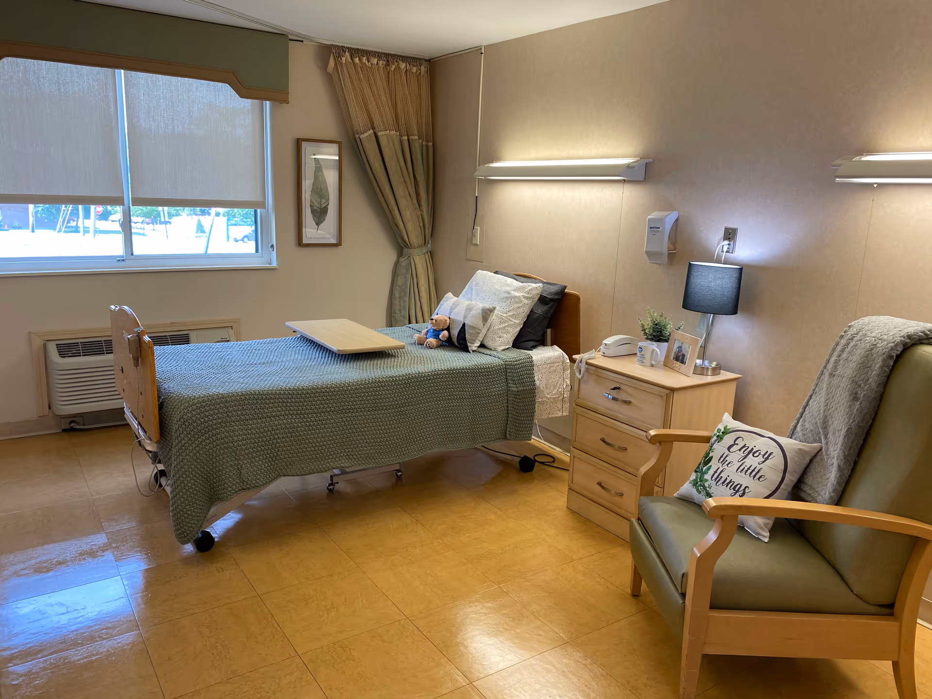Patient bedroom in a senior care facility with a single bed, nightstand, chair, and window blinds.