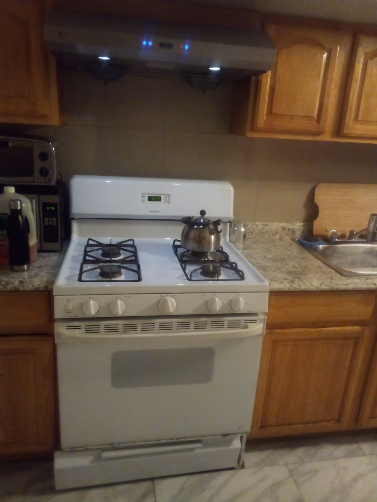 White gas stove with kettle on burner, wooden cabinets, and sink in kitchen.
