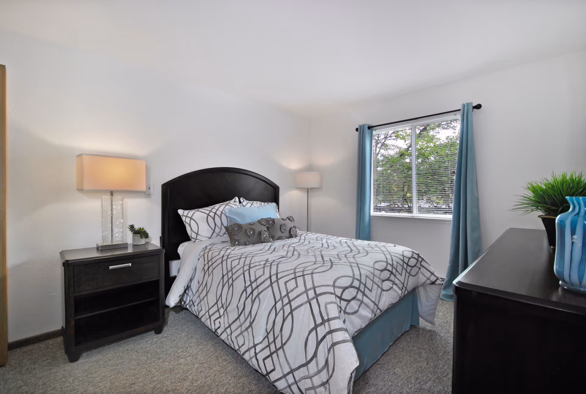 Neatly made bedroom with a dark headboard bed, nightstand, dresser, and a window with blue curtains.