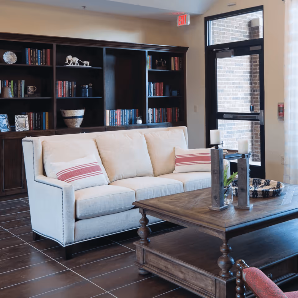A cozy living room area with a beige sofa adorned with two white and red striped pillows, a wooden coffee table with decorative candles and a woven basket, a dark wooden bookshelf filled with books and decorative items, and a glass door leading outside.