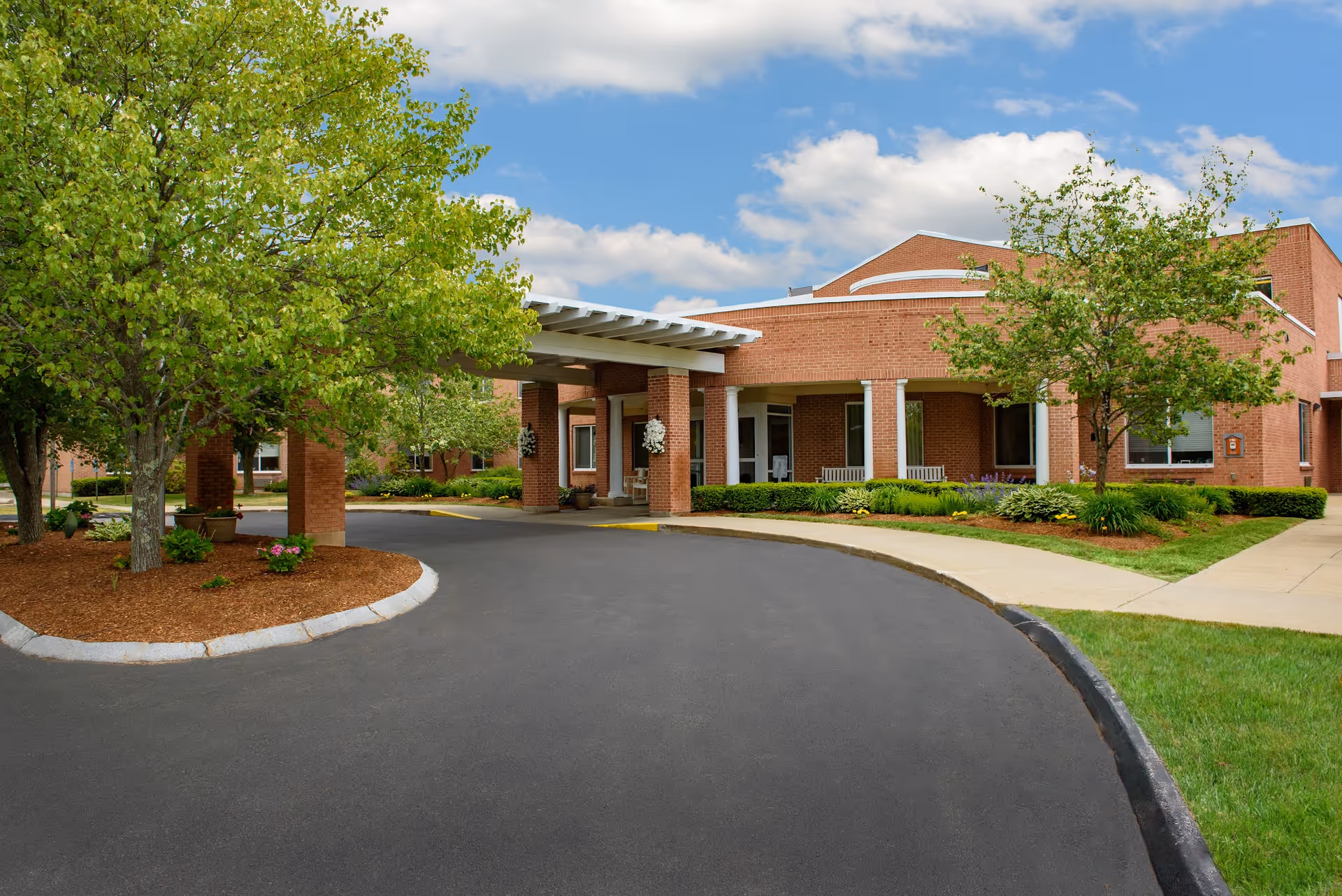 Exterior view of a brick building with a covered entrance driveway, surrounded by green trees and landscaped bushes under a partly cloudy sky.