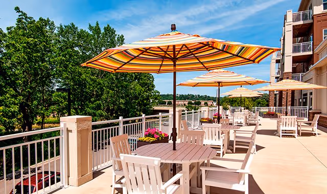 Sunlit outdoor terrace with white tables and chairs under striped umbrellas beside a building, overlooking trees and a blue sky.